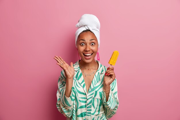 Été, loisirs et dessert froid. Une femme souriante à la peau sombre et positive tient une délicieuse glace à la mangue jaune sur un bâton, se sent excitée et lève la main, porte une serviette enveloppée sur la tête après avoir pris une douche