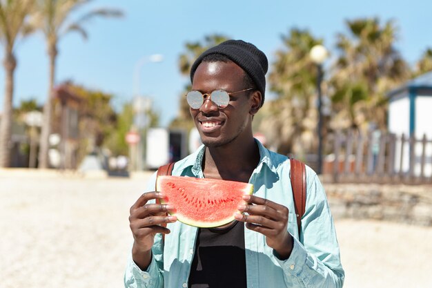 Été, vacances, vacances et style de vie. Heureux jeune voyageur masculin à la peau sombre sans soucis ayant un petit pique-nique avec des amis au bord de la mer, manger de délicieuses pastèques juteuses