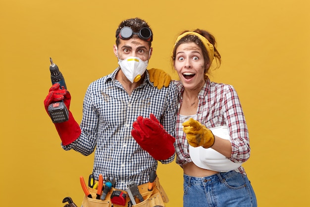 Technicien masculin professionnel portant des lunettes sur la tête, un masque et des gants de protection, une ceinture à outils tenant une perceuse