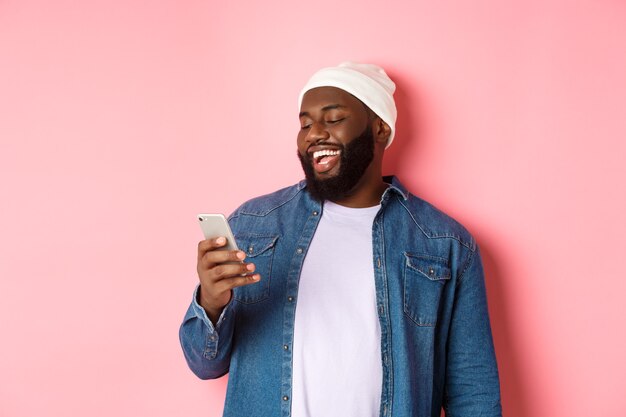 Technologie et concept d'achat en ligne. Heureux homme barbu noir lisant un message et souriant, utilisant un smartphone sur fond rose