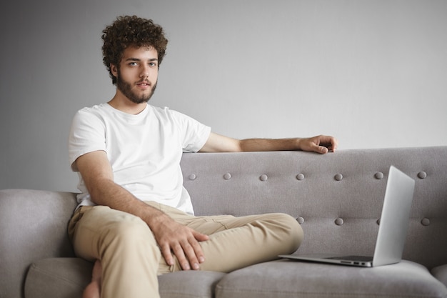 Technologie, gadgets électroniques et concept de communication. Élégant jeune homme aile barbe épaisse et cheveux ondulés assis sur un canapé devant un ordinateur portable ouvert, à l'aide d'une connexion Internet sans fil