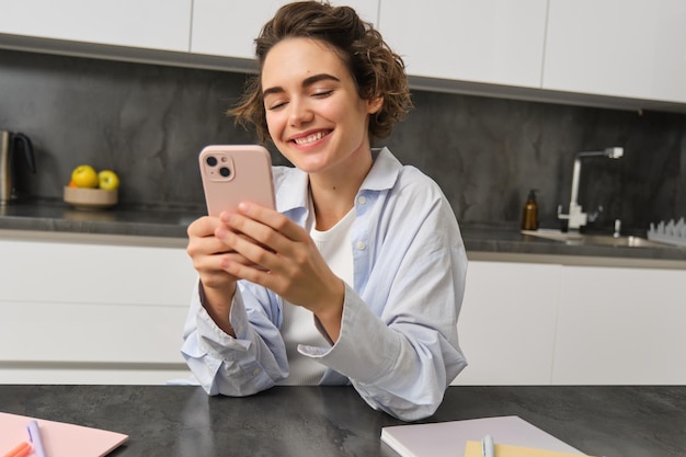 La technologie et le style de vie Une jeune femme est assise à la maison, utilise son smartphone dans sa cuisine et sourit.