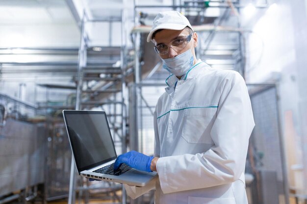 Un technologue en blouse blanche avec un ordinateur portable dans les mains contrôle le processus de production dans la laiterie Lieu d'écriture Le technologue avec un ordinateur portable est à l'usine