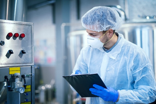 Technologue expert en uniforme de protection avec filet à cheveux et masque prenant les paramètres d'une machine industrielle dans une usine de production alimentaire