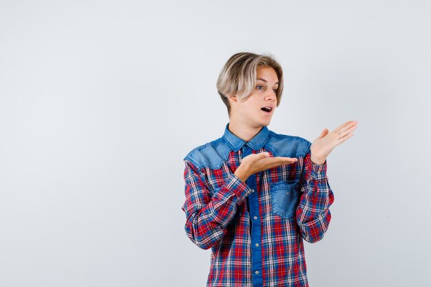 Teen boy faisant semblant de montrer quelque chose en chemise à carreaux et l'air perplexe, vue de face.