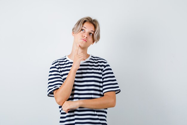 Teen boy in t-shirt gardant le doigt sur le menton et à la pensive , vue de face.