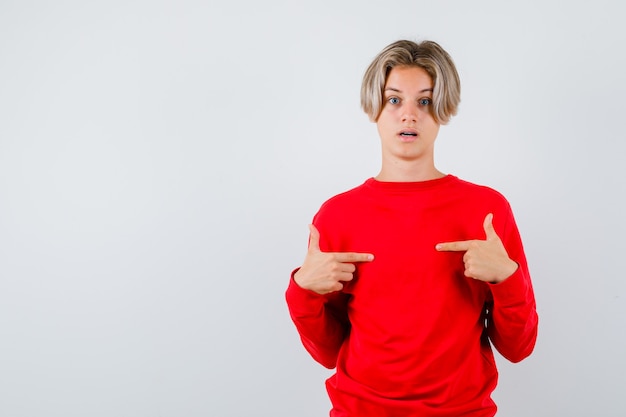 Teen boy pointant sur lui-même en pull rouge et l'air choqué. vue de face.