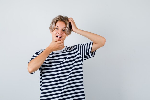 Teen boy se grattant la tête tout en gardant la main sur le menton en t-shirt et l'air étonné. vue de face.