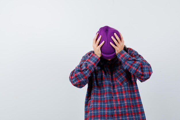 Teen femme en chemise à carreaux et bonnet serrant la tête avec les mains à l'accent