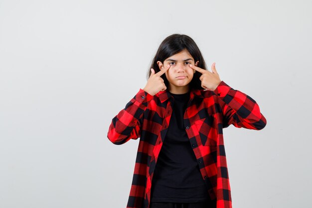 Teen girl holding doigts sur les paupières en t-shirt, chemise à carreaux et à l'imprudente , vue de face.