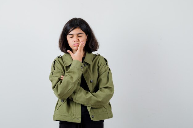 Teen girl holding hand on joue en t-shirt, veste verte et l'air insatisfait. vue de face.