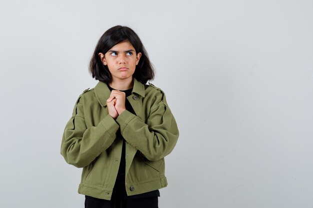 Teen girl holding mains jointes sur la poitrine en t-shirt, veste verte et pensif , vue de face.