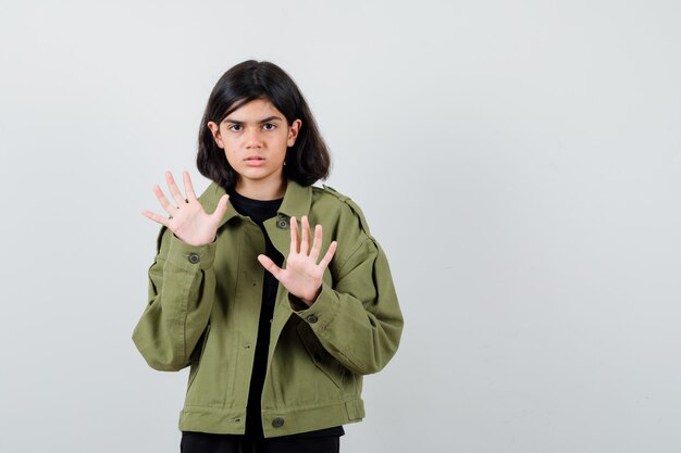 Teen girl in t-shirt, veste verte montrant un geste d'arrêt et l'air confiant, vue de face.