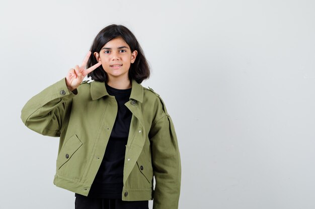 Teen girl in t-shirt, veste verte montrant le signe de la victoire et à la gaieté, vue de face.