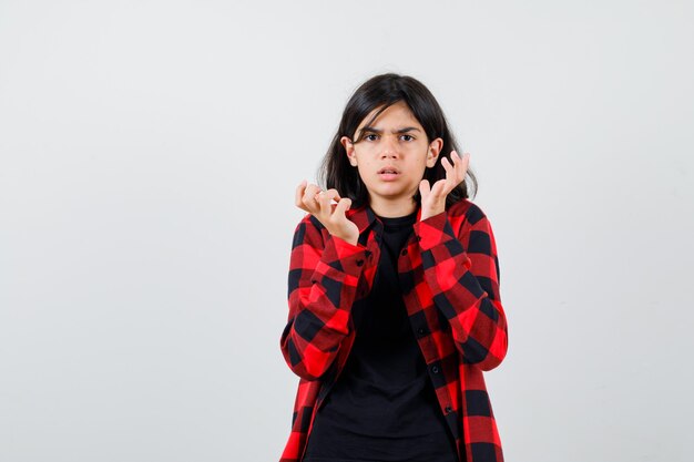 Teen girl levant les mains près du visage en t-shirt, chemise à carreaux et l'air perplexe. vue de face.