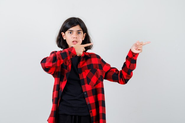 Teen girl pointant vers le côté droit en t-shirt, chemise à carreaux et l'air perplexe. vue de face.