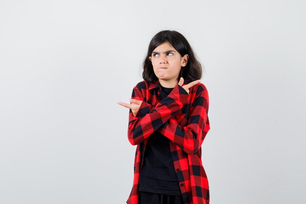 Teen girl pointant vers les deux côtés en t-shirt, chemise à carreaux et l'air indécis. vue de face.