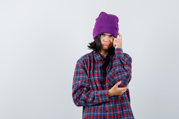 Teen woman holding finger on head en chemise à carreaux et bonnet à pensive