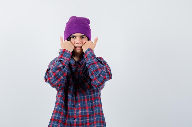 Teen woman holding poings sur les joues en chemise à carreaux et beanie à la prudence
