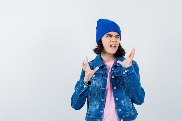 Teen woman in pink t-shirt veste en jean et bonnet levant les mains de manière surprise