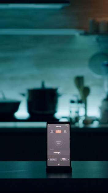 Téléphone avec logiciel intelligent placé sur une table dans la cuisine sans personne à l'intérieur, contrôlant la lumière avec une application de haute technologie