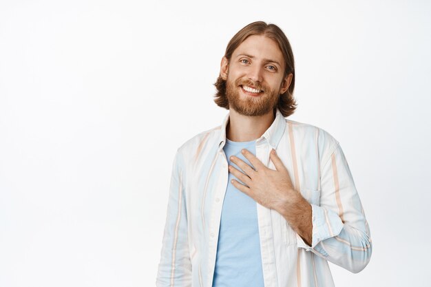 Tellement flatté. Un homme barbu blond souriant tient la main sur le cœur et a l'air heureux, vous apprécie, remercie pour l'aide, exprime sa gratitude, debout sur blanc