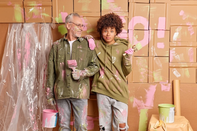 Photo gratuite temps de réparation et concept de travail d'équipe. une femme et un homme de race mixte se tiennent ensemble avec des outils et peignent les murs de la maison occupés à rénover ensemble le salon. deux peintres professionnels