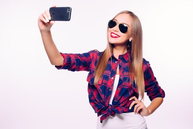 Temps de selfie. Jolie jeune femme en tenue décontractée faisant selfie par son téléphone intelligent et souriant en se tenant debout contre le mur gris