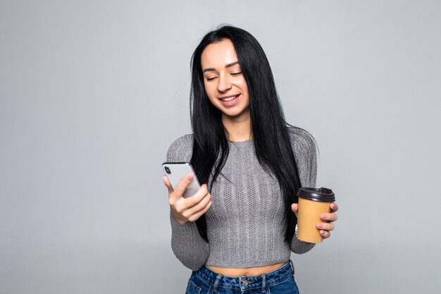 Tendance jeune femme debout discutant sur un téléphone mobile tout en tenant une tasse de café à emporter dans son autre main, isolé sur mur gris