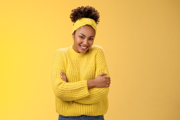 Tender délicate jeune amie afro-américaine joyeuse copine bouclée coiffure bandeau chandail s'embrassant embrassant joyeusement la caméra souriante se sentir douce à l'aise, debout sur fond jaune chaud.