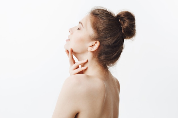 Tendre belle fille européenne aux cheveux noirs en coiffure chignon et type de corps osseux, posant avec les épaules nues, touchant le cou avec les mains, jetant la tête en arrière avec les yeux fermés dans un look détendu et calme.