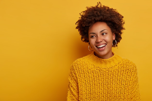 Tendre Charmante Femme Frisée Heureuse A Détendu L'expression Du Visage Joyeux, Coiffure Afro, Porte Un Pull Tricoté, Rit Enthousiaste, Pose Sur Fond Jaune