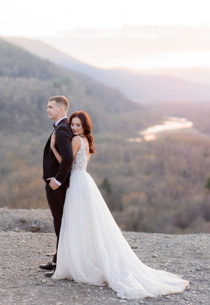 Tendre couple de mariage sur le crépuscule au sommet d'une colline est étreignant, vêtu d'une tenue de mariage de luxe