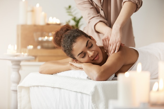 Tendre femme africaine souriante bénéficiant d'un massage avec les yeux fermés dans la station thermale.