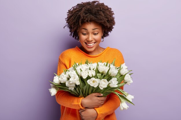 Tendre femme afro-américaine positive embrasse tendrement des fleurs blanches, sourit doucement et regarde les tulipes, porte un pull orange