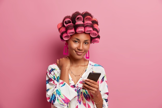 Tendre femme au foyer rétro avec des rouleaux de cheveux, visage de beauté, tient un téléphone portable, regarde la vidéo, vêtue d'une robe de chambre décontractée, pose à l'intérieur.