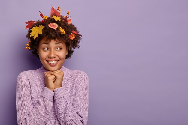 Tendre femme féminine garde les mains sous le menton, exprime des émotions positives après la marche d'automne, a le feuillage dans les cheveux bouclés habillé en pull