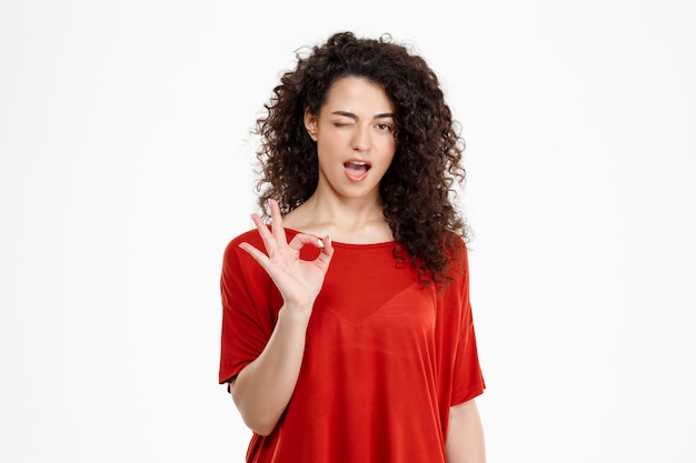 Tendre fille bouclée souriant et pointant signe ok sur mur blanc