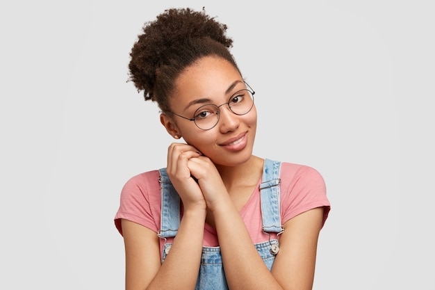 Photo gratuite tendre heureux métis femme avec un sourire charmant, garde les mains près du visage, porte des lunettes incline la tête, porte des vêtements décontractés, isolé sur un mur blanc, heureux d'être invité à une fête avec des amis