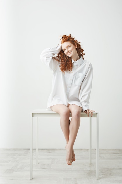 Tendre jeune femme rousse en chemise assise sur la table souriant à corriger les cheveux.