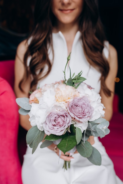 La tendre et jolie mariée est assise avec un riche bouquet de mariage sur un canapé rose au café