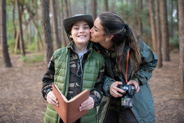 Photo gratuite tendre mère avec fils avec caméra en forêt. modèle féminin en vêtements sportifs embrassant un garçon souriant sur la joue. passe-temps, concept de photographie