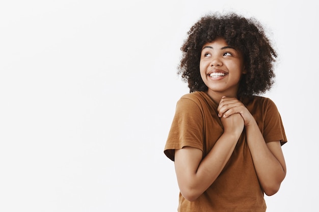 Tendre et rêveuse jolie femme aux cheveux bouclés à la peau foncée en t-shirt tendance marron tenant les mains serrées sur la poitrine et regardant le coin supérieur gauche avec affection