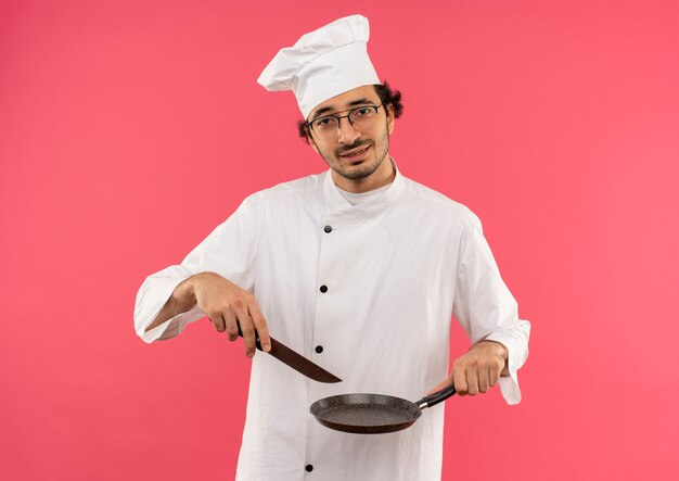 Tendu jeune homme cuisinier portant l'uniforme de chef et des verres tenant une poêle et un couteau