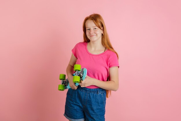 Tenir le patin. Portrait d'une adolescente caucasienne sur fond de studio rose corail. Beau modèle rousse féminin. Concept d'émotions humaines, expression faciale, ventes, publicité, culture des jeunes. Espace de copie.