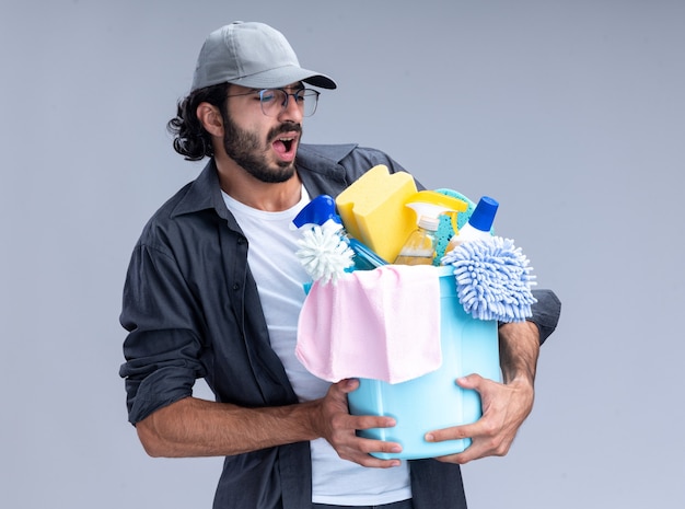 Tense jeune beau mec de nettoyage portant un t-shirt et une casquette tenant un seau d'outils de nettoyage isolé sur un mur blanc