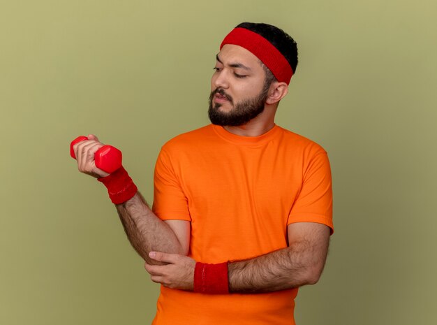 Tense jeune homme sportif portant bandeau et bracelet exercice avec haltère isolé sur fond vert olive