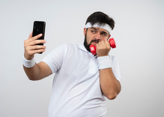 Tense jeune homme sportif portant bandeau et bracelet exercice avec haltère prendre un selfie isolé sur un mur blanc