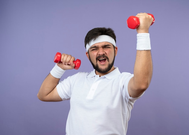 Tense jeune homme sportif portant bandeau et bracelet exercice avec des haltères isolé sur mur vert