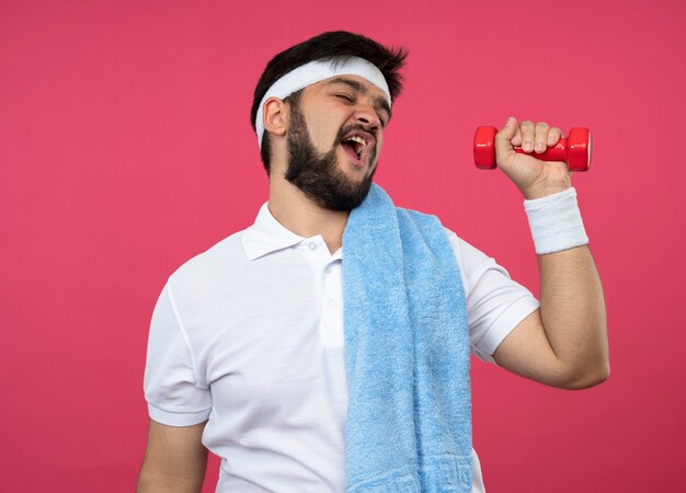 Photo gratuite tense jeune homme sportif portant bandeau et bracelet avec une serviette sur l'épaule exercice avec haltère isolé sur rose